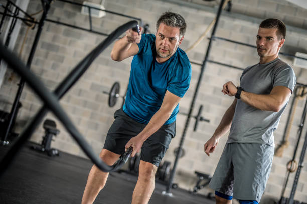 Mature man using battling ropes in gym under supervision of personal trainer.
